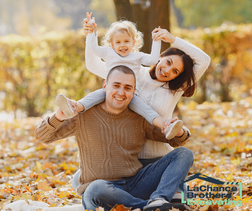 A man kneels on fallen autumn leaves while lifting a child on his shoulders; a woman sits beside them, smiling, with a child in her arms in a park with golden foliage.